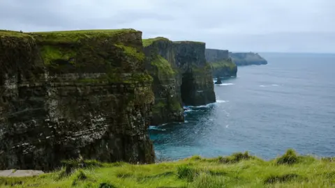 Getty Images A view of the Cliffs of Moher on a cloudy day with a calm sea below. The tall, rugged cliffs jut out at angles to the sea.