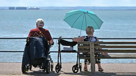 Getty Images A man and woman sit in the sun overlooking the sea. She shelters under a blue umbrella.