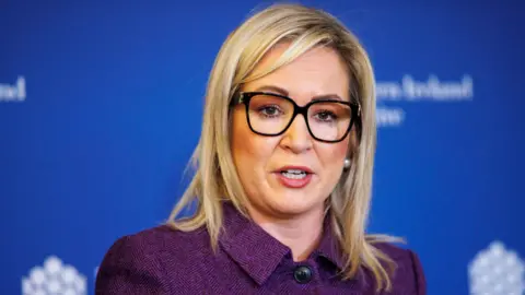 PA Media Michelle O'Neill, with shoulder-length blonde hair. She is wearing a purple jacket and glasses with a black frame. There is a blue wall behind her. 