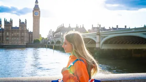 Getty Images A young woman wearing a bright orange jacket and a Union Jack-coloured bag on her shoulder walks along the bank of the river Thames. Big Ben and the Houses of Parliament is in the background behind her.