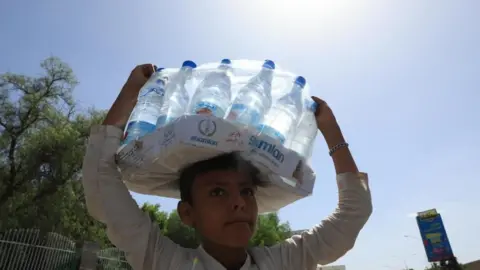 EPA A boy carries a box of bottled water on his head. He is wearing a white-coloured shirt. There is bright sunshine in a blue sky behind him, as well as trees and a road sign. 