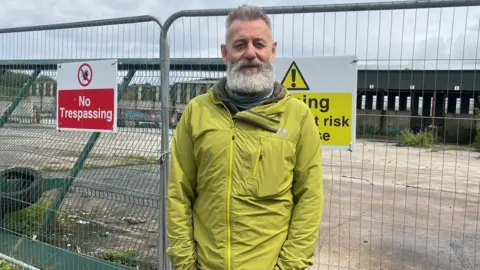BBC A man is standing in front of a metal fence. He has a beard and is wearing a green coat. No trespassing signs are on the fence.