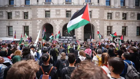 PA Media A crowd of protesters standing and looking at the Foreign Office building, holding Palestinian flags and a sign that reads: Stop Israel starving the 2.3 million in Gaza