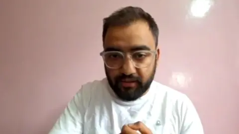 BBC A man wearing a white t-shirt with spectacles looks into the camera.
