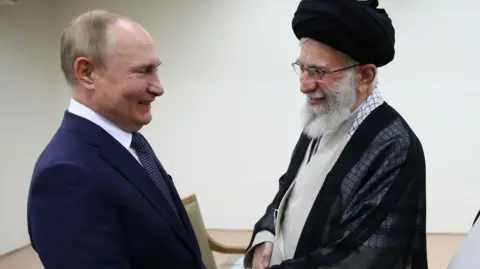 Reuters Putin on the left shakes the hand of Ayatollah Khamenei. Both men are smiling. Putin is wearing a navy suit while Khamenei is wearing traditional Islamic clerical robes and a black turban, along with glasses.