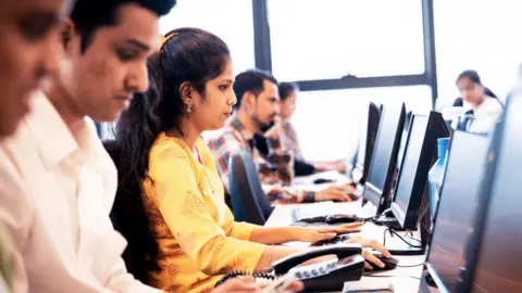 Getty Images The image shows Indian workers at a software company seated in front of their computer screens. A woman wearing a white kurta is prominently seen, surrounded by male colleagues on either side. 