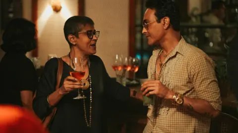 Soho House A man and a woman at a club having a drink and engaged in an animated chat. The man is wearing a checked shirt with sunglasses while the woman is wearing a black dress with glasses.
