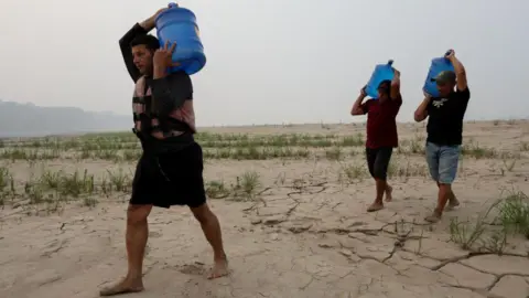 Getty Images Three men carry drinking water above their heads in large blue plastic tanks along a dry sandbank of the Madeira River in northern Brazil
