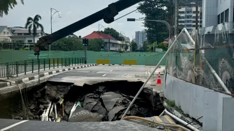 PUB A deep sinkhole in the middle of a road in eastern Singapore. The road is fenced off from traffic and a crane is hovering above the sinkhole. Residential buildings and shophouses are seen in the background