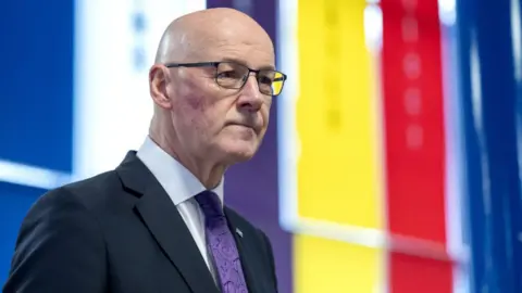 PA Media A bald man with glasses stands in front of a striped multi-coloured background. He is wearing a dark suit, white shirt and purple tie.