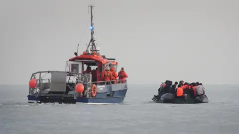 PA Media A French rescue boat with crew members in orange uniforms and life jackets escorts a small, overcrowded inflatable boat. The inflatable boat is filled with people wearing life jackets, believed to be migrants, as it moves away from the French coast.