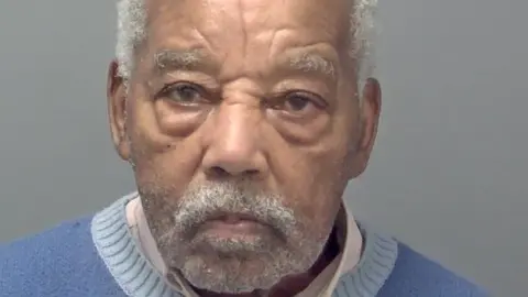 Avon and Somerset Police Ryland Headley's custody picture. He has short white hair, and short facial hair, and wears a white collared shirt under a blue jumper. He is looking straight ahead with a serious expression.