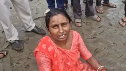 ANI A woman wearing an orange saree sitting on wet sand with the feet of people standing behind in the frame 