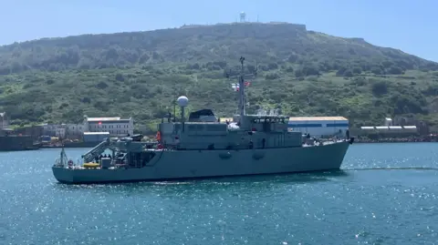 BBC A warship on the water sailing to the right with a union jack visible and a Ukraine Navy flag also visible with some green hills in the background