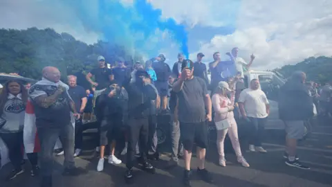 PA Media A group of mostly male protesters, some standing on the back of flatbed truck with a white cab. Some are wearing caps or face coverings, while others point their mobile phone cameras towards the photographer. The air is smoky from flares and plumes of blue smoke are rising up from the crowd
