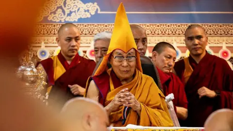Getty Images The Dalai Lama in a saffron robe and conical hat, sitting on a chair with his hands clasped together. Behind him are other monks in robes.