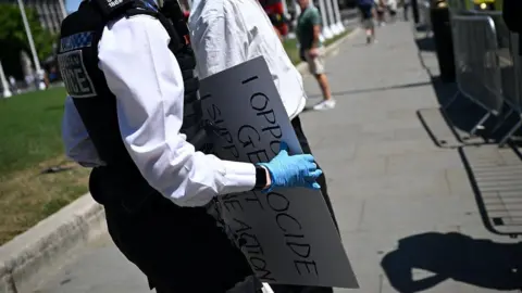 AFP via Getty Images A close up shot of a female police officer's torso as she holds a sign that she has confiscated that reads: I oppose genocide, I support Palestine Action. She is wearing police uniform and blue gloves