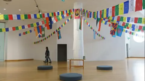 Constellation of Complicity A large pillar stands in the middle of a gallery wrapped in white cloth, with several strings of colourful bunting carrying the flags of various countries and Tibetan art tied to the top of the pillar where a red and yellow flag has been pinned. There is a small wooden table at the base of the pillar as well as black circular cushions placed around it. Two people dressed in jeans and dark T-shirts are walking across the gallery