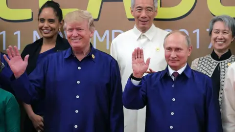 Getty Images Donald Trump and Vladimir Putin hold their hands up in a wave while wearing matching blue overshirts, as they pose with other leaders for a "family photo" in Vietnam in November 2017