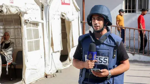 AFP via Getty Images Anas al-Sharif stands next to a tent wearing a dark blue helmet and vest with "press" written on it, while he speaks into a microphone during a broadcast.