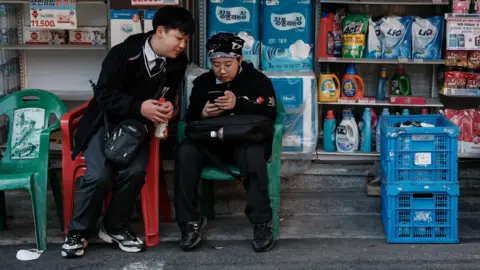 Getty Images A South Korean schoolboy, wearing a school uniform and glasses, scrolls on his phone while sitting in a plastic chair outside a convenience store. Another boy in the same uniform leans over from his own chair to watch the screen.