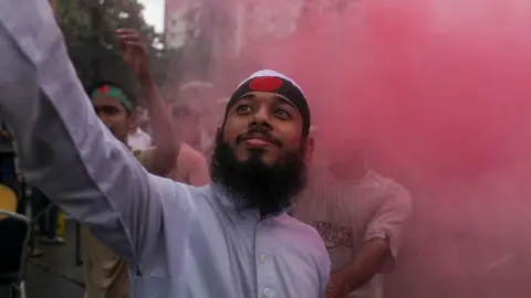 NurPhoto via Getty Images A bearded young man in a blue shirt stands waving a can making red smoke in amongst a crowd, wearing a green headband with a red dot in the middle -  a version of the national flag. Blurry people in a crowd can be seen in the background