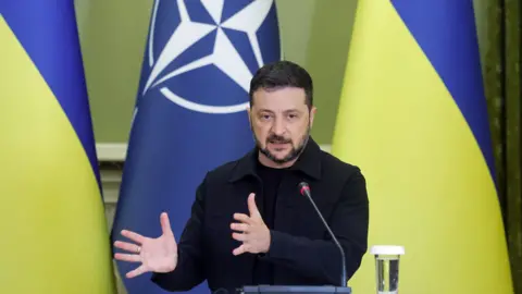 EPA/Shutterstock Ukrainian President Volodymyr Zelensky, dressed in his signature black, gesticulates as he addresses a joint press conference in Kyiv. Behind him his the Ukrainian flag.