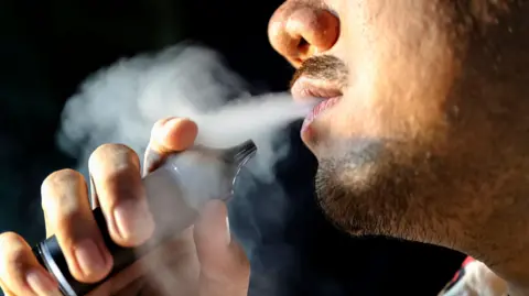 Getty Images A close-up of the lower half of a man's face as he exhales a white cloud of vape smoke, while he clutches a black coloured vape close to his face