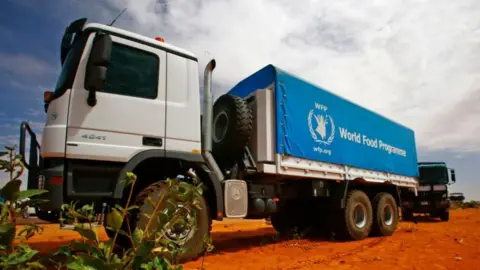 AFP via Getty Images An aid lorry with a blue tarpaulin that says World Food Programme is travelling on a red dusty road - two other vehicles can be seen behind it on the same road. 