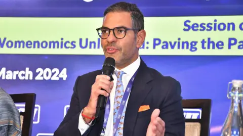 Sunjay Kapur/X Sunjay Kapur wearing a black suit with a blue printed tie and a white shirt holding the mic. 