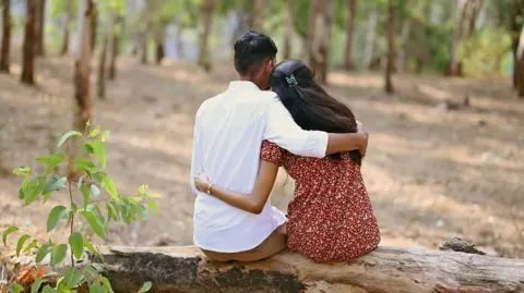Getty Images A boy and girl put their arms around each other in a garden in India. The boy is wearing a white shirt while the girl is wearing a maroon tunic with pattern.