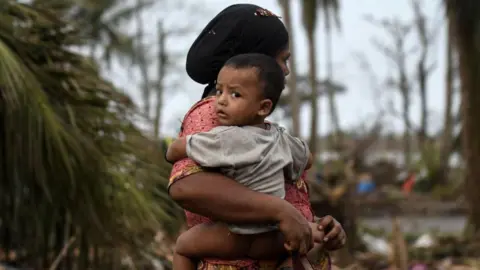 Getty Images Representational Image: A Rohingya woman carries her baby next to her destroyed house at Basara refugee camp in Sittwe on May 16, 2023, after cyclone Mocha made a landfall.