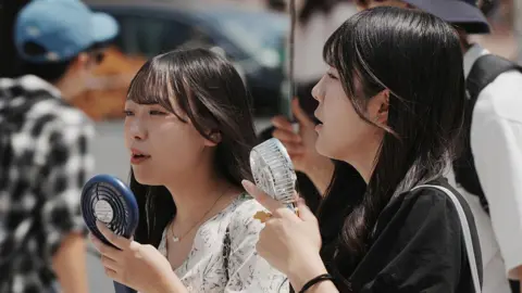 Getty Images Two girls with long black hair holding mini fans to their faces as they stand on a road.