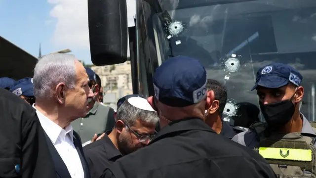 Netanyahu, surrounded by people, stares at a bus windscreen which has bullet holes in