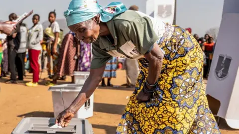 Reuters A woman in a head-scarf and patterned yellow wrap places a vote in a ballot box. The box is on a dusty field at an outdoor polling station - a crowd of people waiting to vote can be seen in the background.