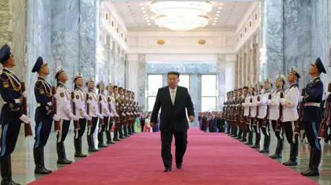 KCNA via EPA North Korean leader Kim Jong Un arriving to attend a ceremony in Pyongyang.  He is wearing a black suit and walking down a red carpet, between two rows of guards holding rifles, dressed in formal uniforms. Above him are big white chandeliers. 