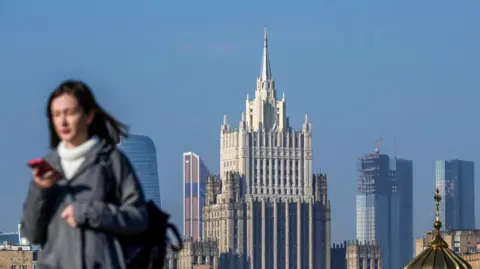 NATALIA KOLESNIKOVA/AFP via Getty Images Photograph showing a woman looking at her phone as she walks across a bridge in central Moscow - with the Russian Foreign Ministry building in the background