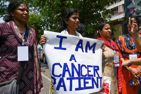 Hindustan Times via Getty Images A woman who is a cancer survivor and a teacher participating in a demonstration in Kolkata in April 2025, holding a "I am a cancer patient" placard. 
