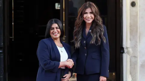 Reuters Home Secretary Shabana Mahmood shakes hands with U.S. Homeland Security Secretary Kristi Noem . Mahmood and Noem are both smiling and wearing navy blue suit jackets.