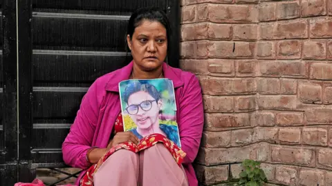 Pritam Roy A woman sits holding a picture of her loved one. She is dressed in a bright pink top and pale pink trousers