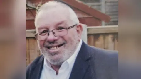 BBC Melvin Cravitz smiles. He is wearing a dark kippah hat, glasses and a dark suit jacket with a white shirt.