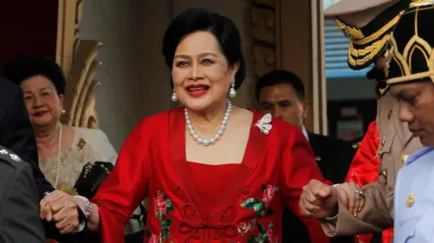 Reuters Queen Sirikit, wearing pearl jewellery and a red dress, is helped down a staircase by guards in uniform.