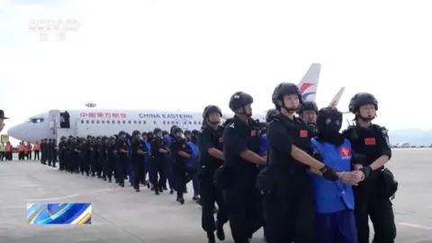 CCTV Rows of security personnel in black clothes and helmets escorting suspects from a plane. Each suspect, wearing bright blue attire and a black mask over their heads, is flanked by two guards.