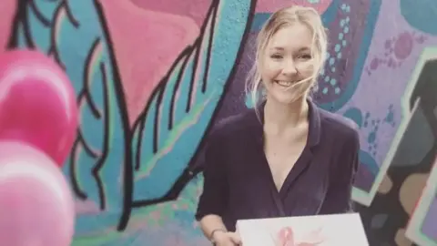 Supplied A blonde young woman smiles at the camera holding a white box tied with a ribbon. Behind her is a colourful mural