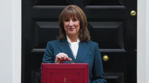Getty Images The chancellor, Rachel Reeves, wears a blue suit and white shirt as she holds up her red box in front of the black door to No11 Downing Street.