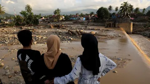 Reuters Two women in Muslim headscarves and a man look at muddy ground strewn with stones.  