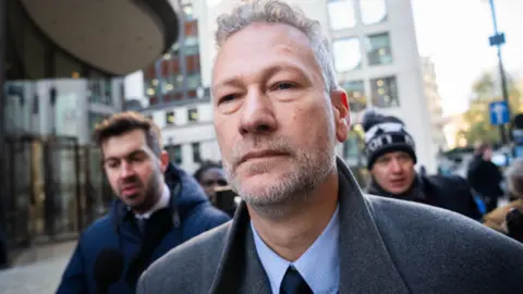 James Manning/PA Wire A close-up photo of Nathan Gill as he arrives at court. He has short grey hair and a closely-shaven beard; he wears a grey winter coat with a light blue shirt and dark blue tie. There are people behind him, to his left and right, including a man with a microphone.