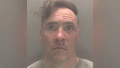 CPS A mugshot of Paul Doyle wearing a grew jumper looking stunned. His hair, a long brown fringe, is dishevelled and to the side. He is wearing a grey t-shirt.