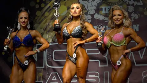 BBC Roya Karimi (centre) on stage holding up her trophy, alongside the silver and bronze winners in the Wellness category of the Stoperiet Open bodybuilding competition