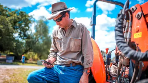 Getty Images A farmer wearing a hat, sunglasses, jeans and shirt leans on a tractor and looks at a smartphone.
